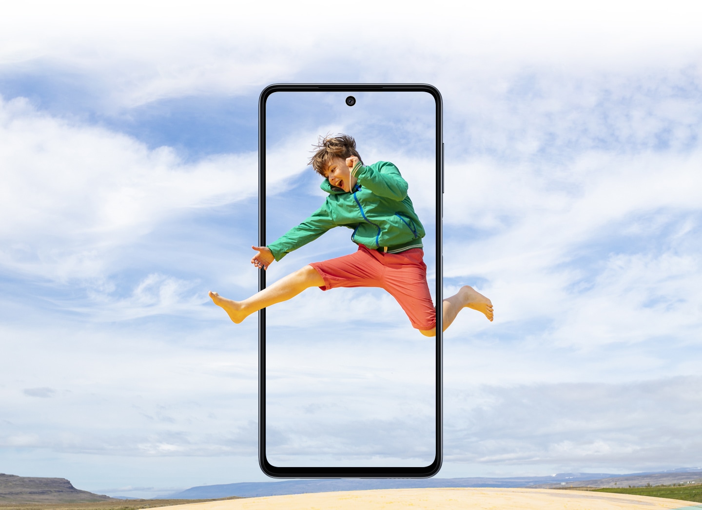 Galaxy M52 5G seen from the front. A playful girl jumping up in motion captured in a vertical M52 5G display and the big blue sky expands outside of the boundaries of the display.