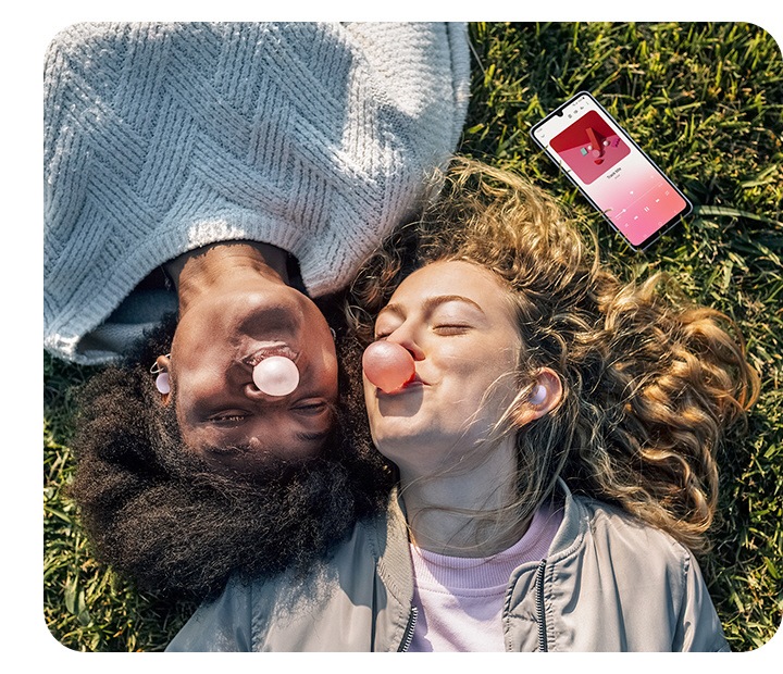 Two female friends, both wearing Buds Pro, are lying down on the grass on a sunny day, blowing bubbles with bubble gum. A smartphone laid down nearby shows a song being played, illustrating that both friends are listening to the song together with their Buds products.