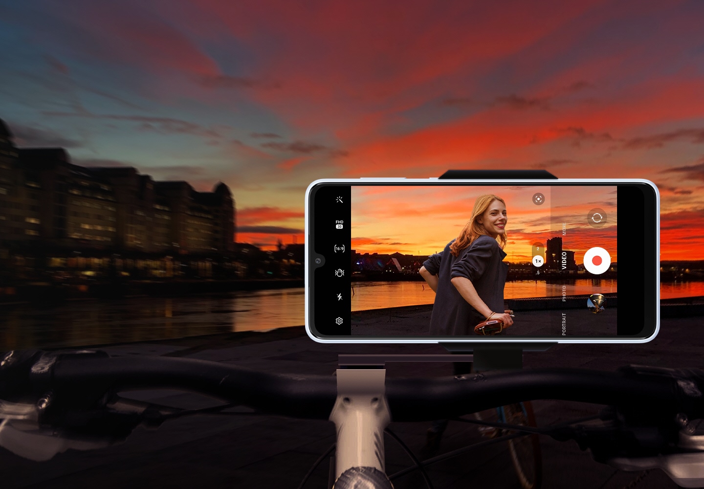 A Galaxy A33 5G is mounted in landscape mode on top of a bicycle handlebar. In the background, a dark and blurry sunset landscape view is shown. On screen, the same landscape is shown more vibrantly with a woman also captured clearly and without blur.