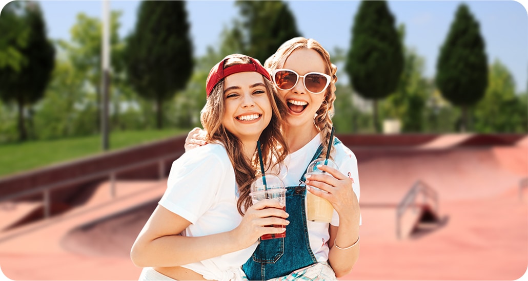 live focus on activated, Two female friends are smiling, looking into the camera in front of a skatepark with trees and grass in the background. With the Live Focus On icon above the shot activated, the background is blurry.