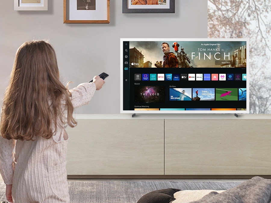 A girl is pointing the remote towards The Frame, which shows the Smart Hub home screen. 