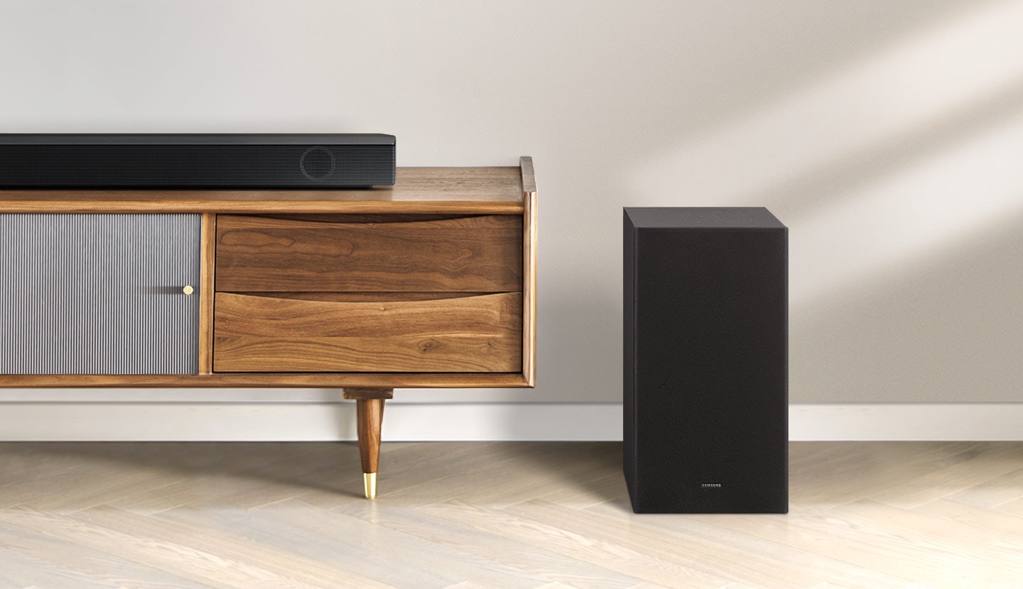 Samsung B series soundbar is being shown on top of a contemporary TV cabinet along with its matching subwoofer which is being displayed to the side of the TV cabinet.