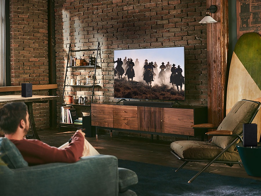 A man enjoys racing content on his TV. Samsung Wireless Rear Speaker Kit and Soundbar create Immersive surround sound. Toggle button is shown.