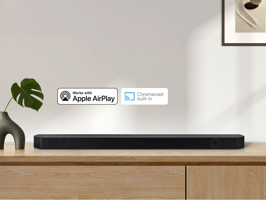Apple AirPlay logo, Chromecast Built-in logo, and Ok Google logo can be seen along with Samsung Q990B soundbar which is sitting on living room cabinet.