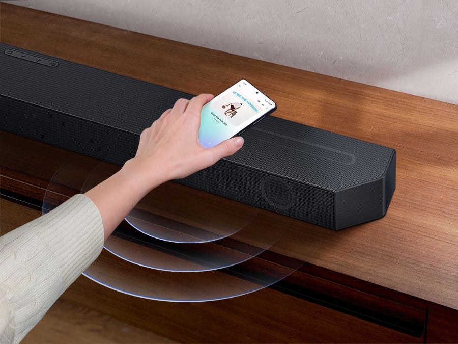 A hand taps a smartphone with the Samsung music app on-screen on the Soundbar. It instantly plays music, showing how easy it is to switch from smartphone to Soundbar.