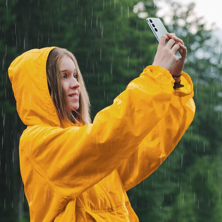 A smiling person in a bright yellow raincoat taking a selfie with a Galaxy A55 5G in the rain.