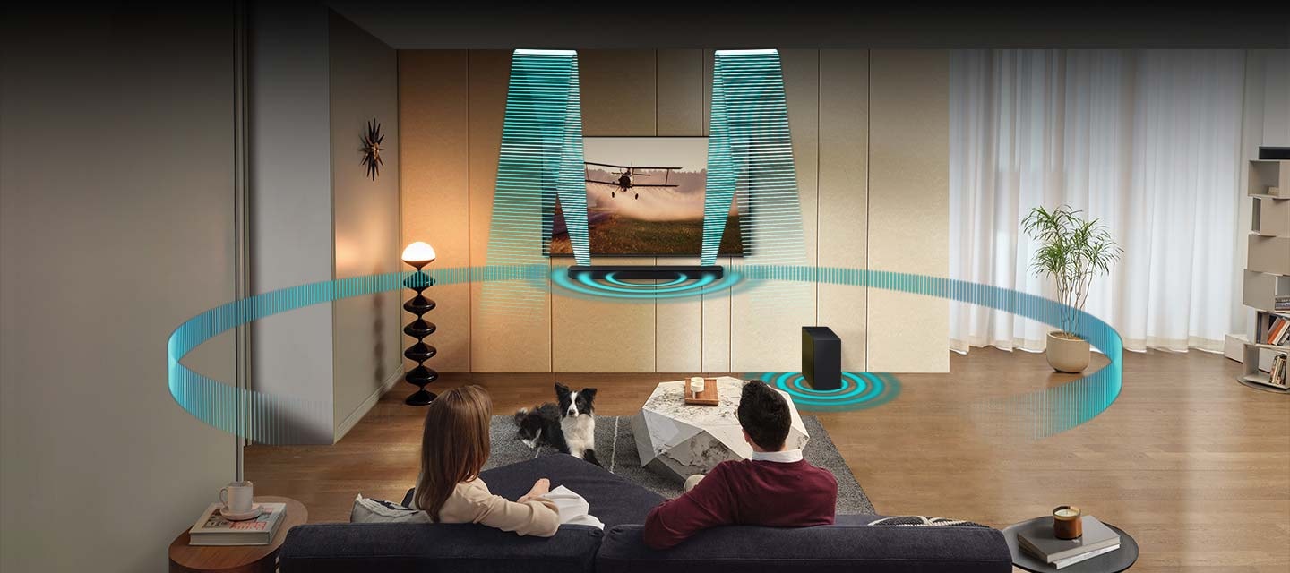 In a living room, a couple sits on a sofa and watches TV. The TV is installed onto the wall, with a Soundbar and subwoofer underneath. Sound waves emanate from the Soundbar in various angles, traveling in multiple directions to fill the room. Then round sound waves emanate from both the Soundbar and subwoofer.