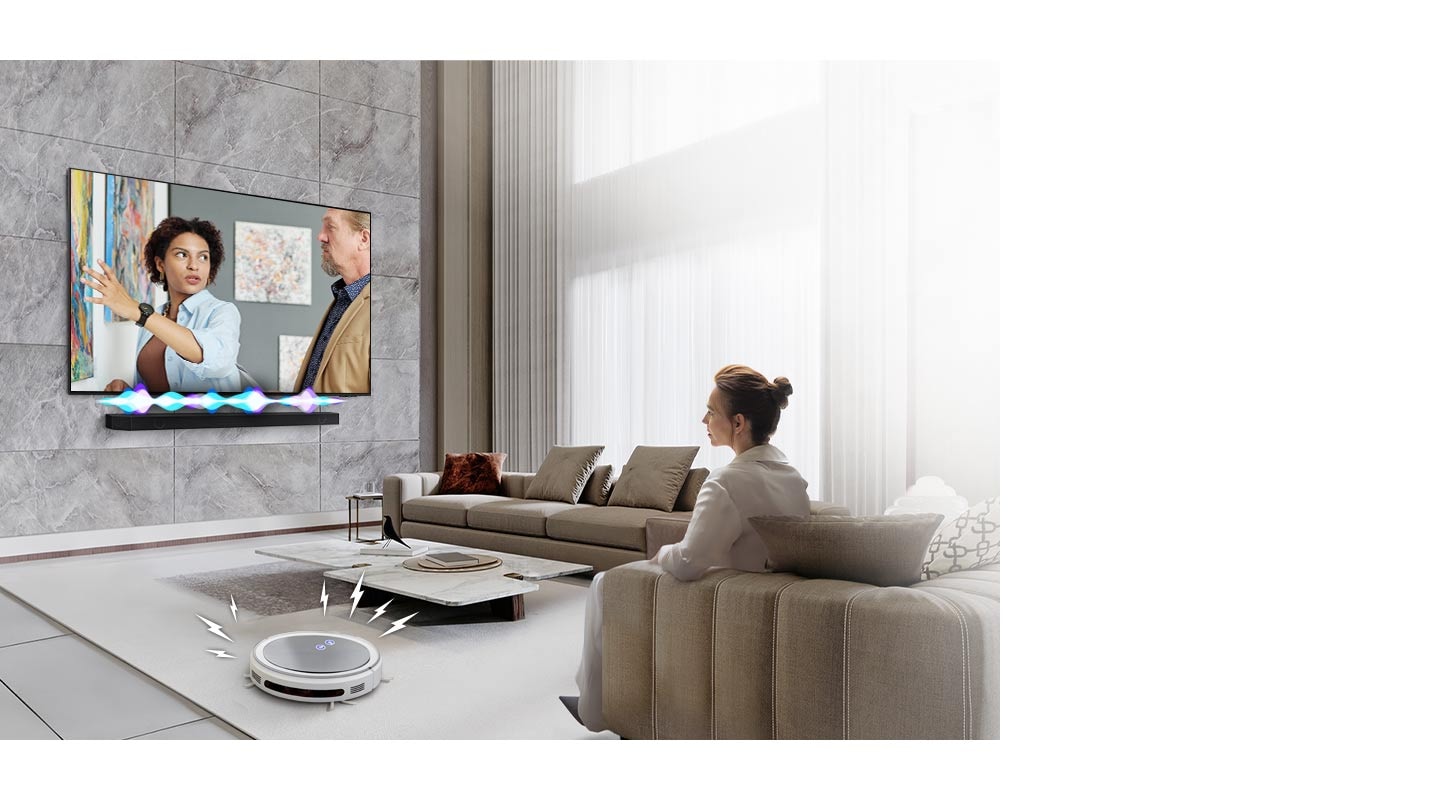 A woman is watching TV, which has a Soundbar installed directly underneath. The Soundbar has an audio bar indicating the level of volume. A robot vacuum cleaner rolls into the room, giving off distracting noise indicated by lighting bolts. In response, the Soundbar plays at a louder volume, with the audio bar giving off greater activity.