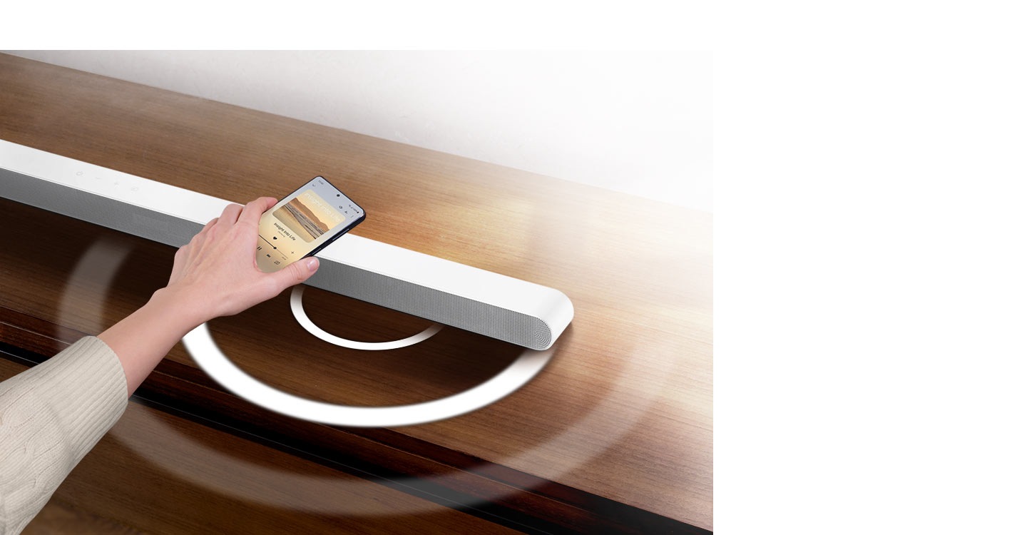 A hand holds a phone playing music and taps it lightly against an Ultra Slim Soundbar. Instantly, the Soundbar gives off a series of round sound waves, indicating that it's playing the same music as the phone.