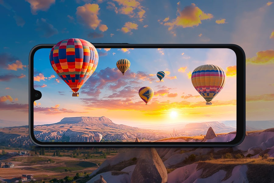 In the background, a canyon landscape at sunset with colorful hot-air balloons floating in the sky. In the foreground, a Galaxy A16 5G device displays its FHD+ Super AMOLED screen, bringing the landscape to life with clearer details and more vivid colors. One balloon appears to float out of the screen, highlighting the immersive viewing experience.