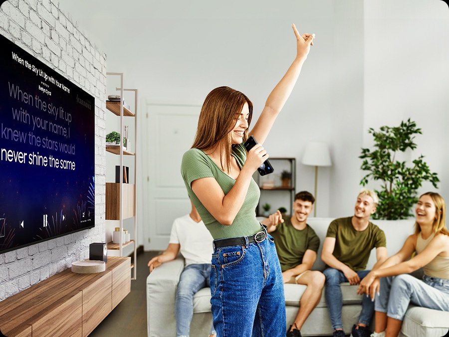 A woman sings into her phone, while the TV behind her displays karaoke lyrics. Her friends smile in the background.