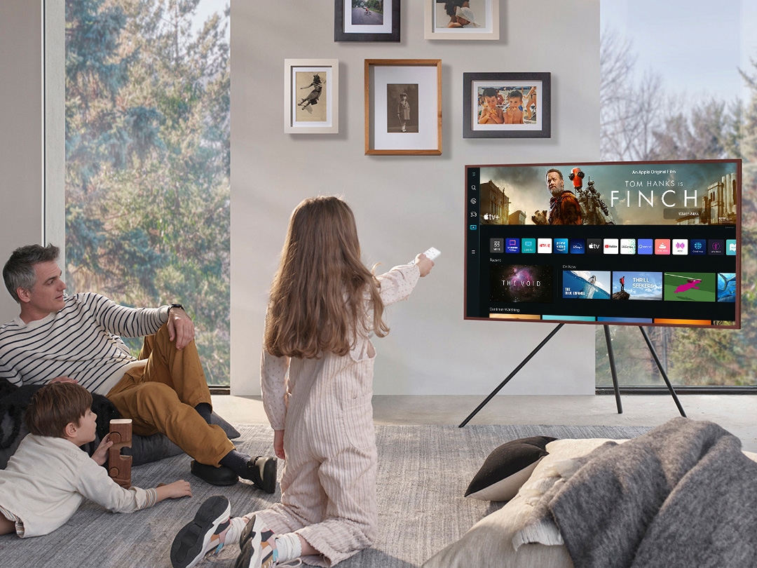 A girl is pointing the remote towards The Frame, which shows the Smart Hub home screen.