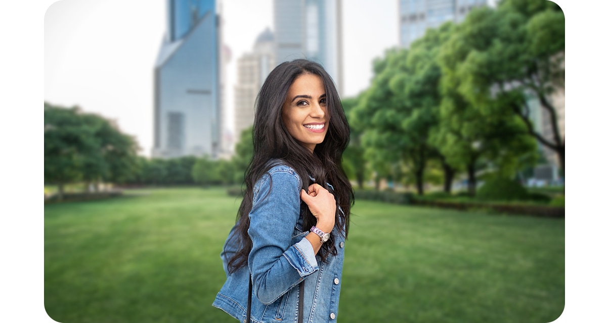 Portrait mode is on. A woman is standing in an urban park, smiling at the camera. The background, which includes grass, trees, and high rise buildings, is blurred slightly so she stands out.
