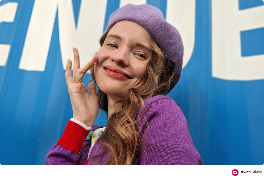 A woman, smiling and wearing a purple outfit, takes a high-quality selfie in front of a blue background. Camera icon and #WithGalaxy text are shown.