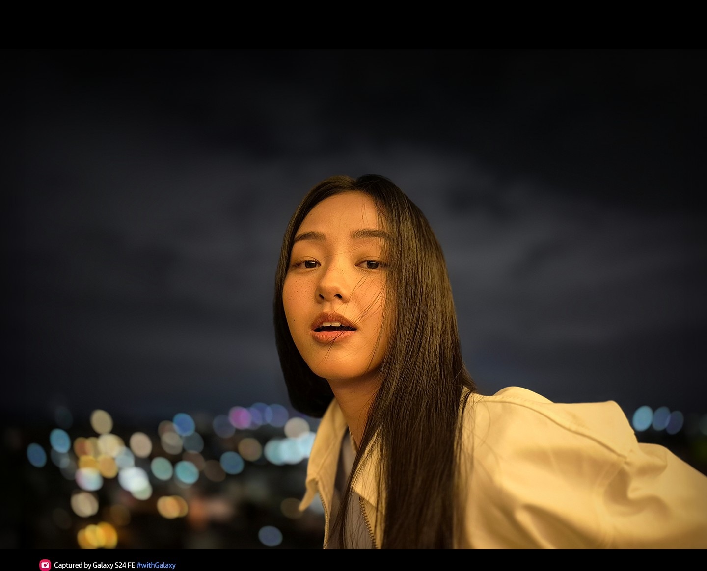 A photo of a woman outdoors at night with a lit face and blurred city lights in the background. Text reads Captured by Galaxy S24 FE #withGalaxy.