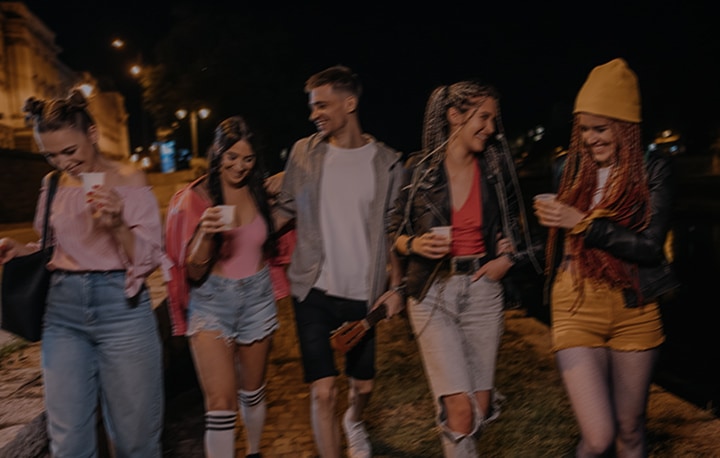 Five friends, four women and one man, walk beside a river at night holding drinks under streetlights. A vertical slider splits the image, with the left side dim and muted and the right side bright and colorful, revealing clearer details like a yellow beanie, ripped jeans, and reflections on the water.