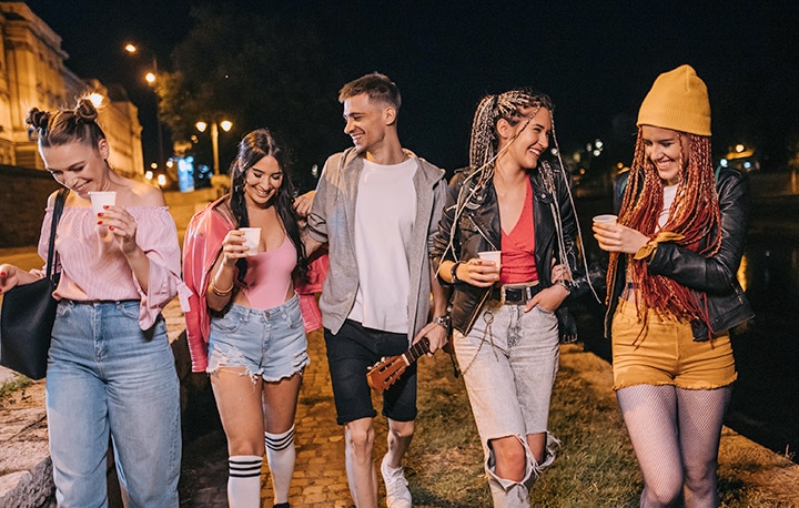 Five friends, four women and one man, walk beside a river at night holding drinks under streetlights. A vertical slider splits the image, with the left side dim and muted and the right side bright and colorful, revealing clearer details like a yellow beanie, ripped jeans, and reflections on the water.