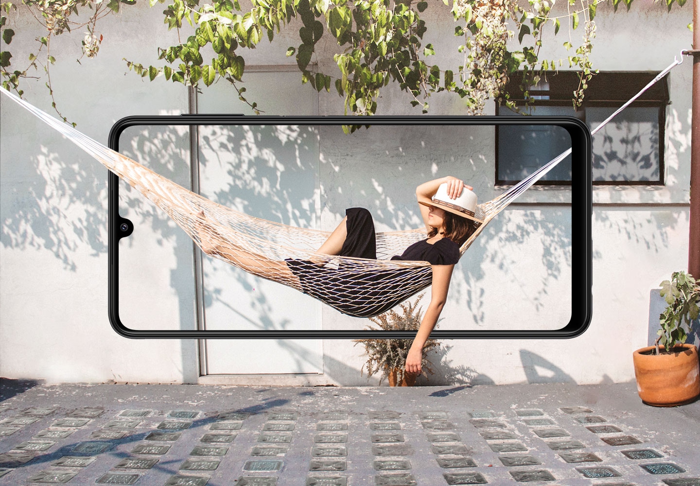 A Galaxy A22 is seen from the front, horizontally. Onscreen, a woman rests in a hammock, which blends beyond the bezel with the side of a house.