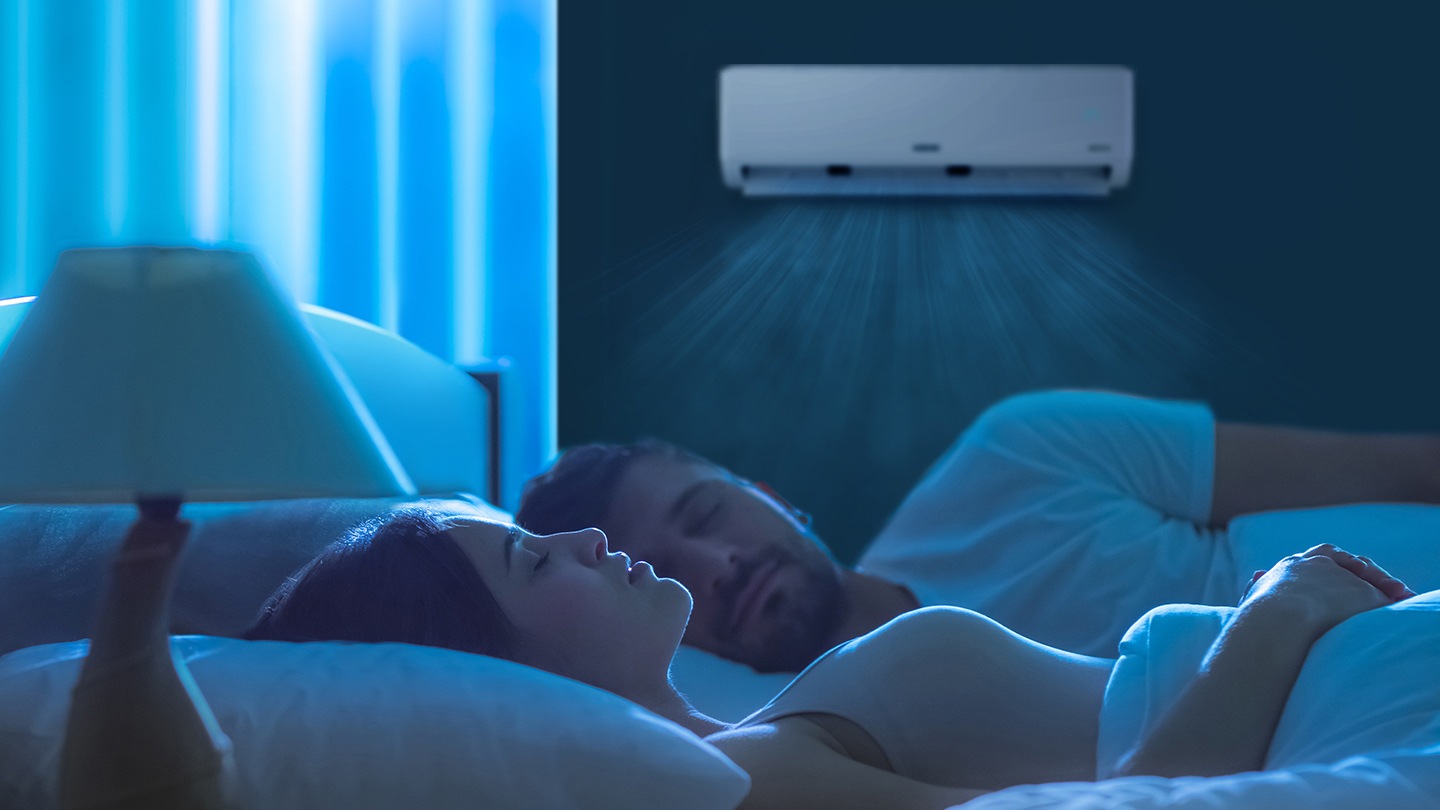 Shows a couple sleeping in bed while a wall-mounted air conditioner quietly creates a comfortable bedroom environment.