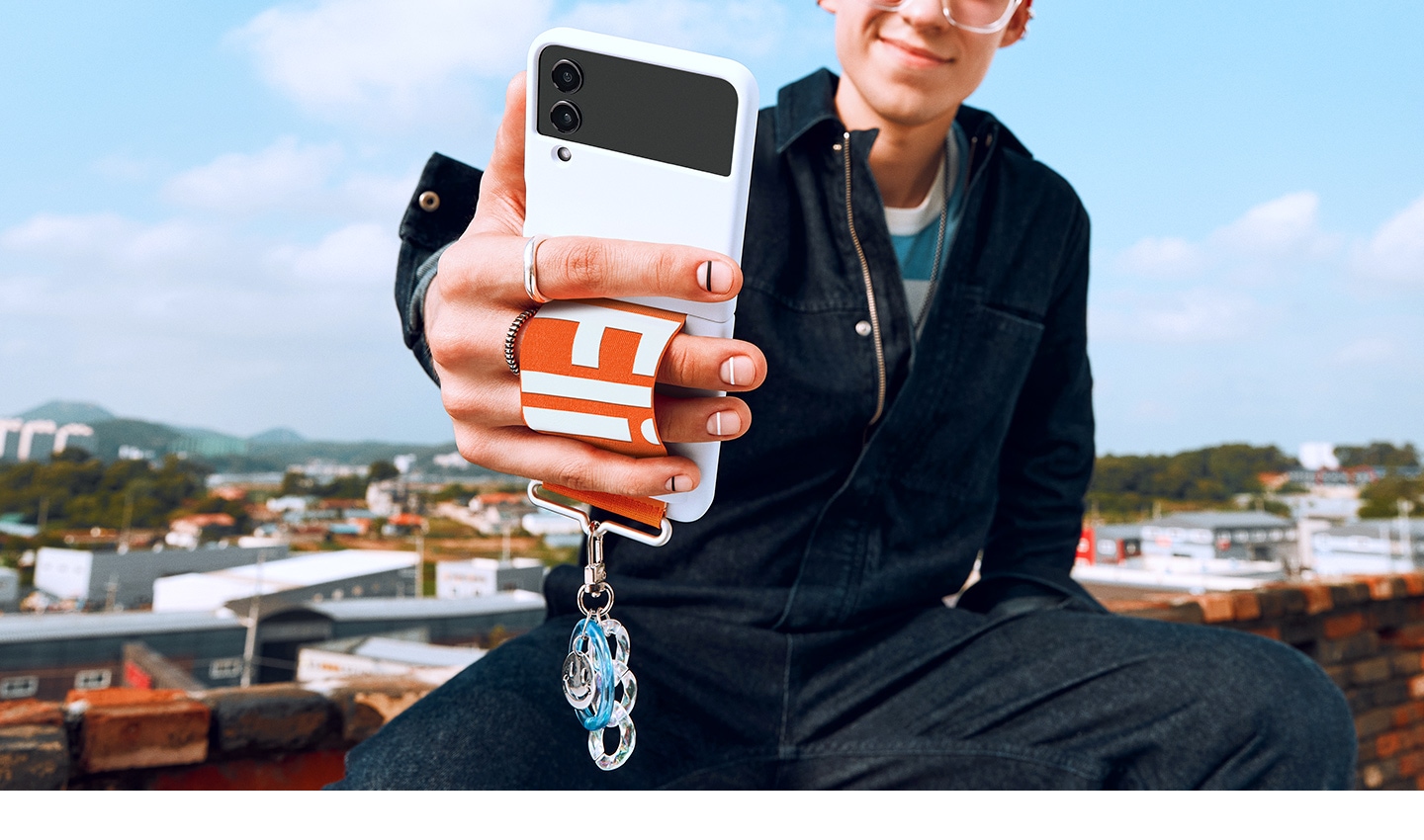Ein lächelnder Mann hält sein Galaxy Z Flip4 nach vorne, um ein Selfie aufzunehmen. Das Galaxy Z Flip4 steckt in einem Silicone Cover with Strap in White, das mit einem orangefarbenem Band ausgestattet ist.