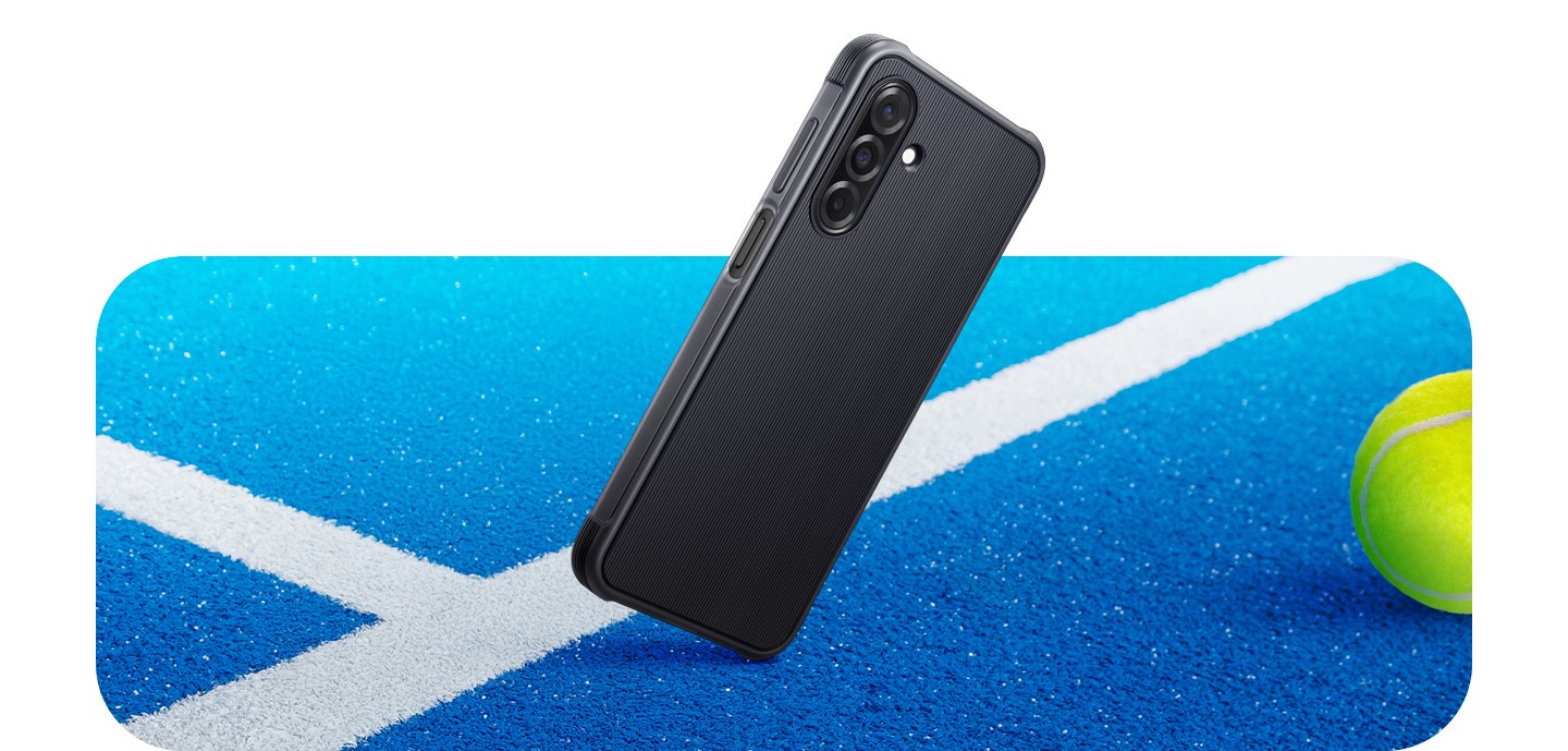 Ein Galaxy A26 5G mit einem angebrachten Rugged Case in Kontakt mit der Oberfläche eines Tennisplatzes, als wäre es auf eine Ecke gefallen.