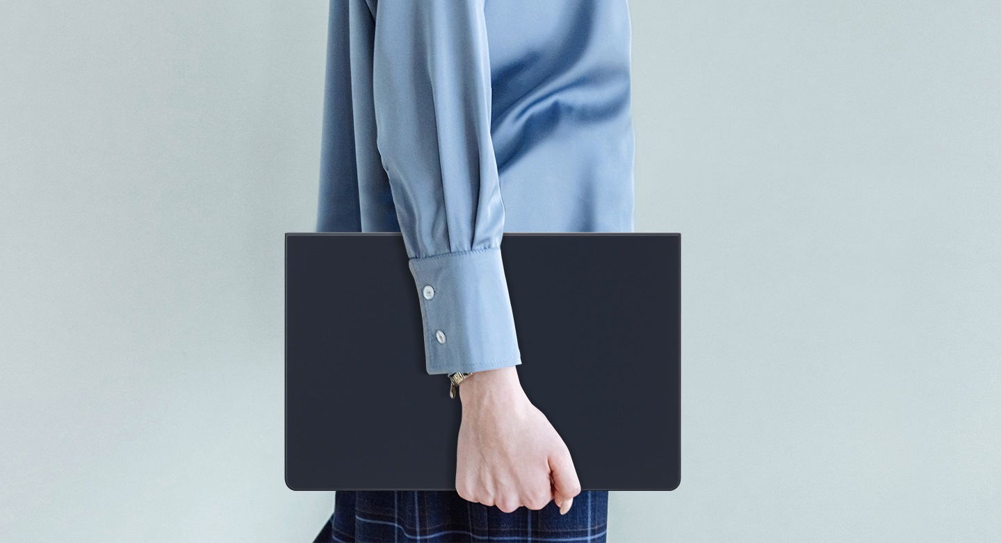Egy kék inget viselő személy sétál, miközben egy Galaxy Tab S9+ készüléket tart, amelyet Book Cover Keyboard Slim borítóval borított.