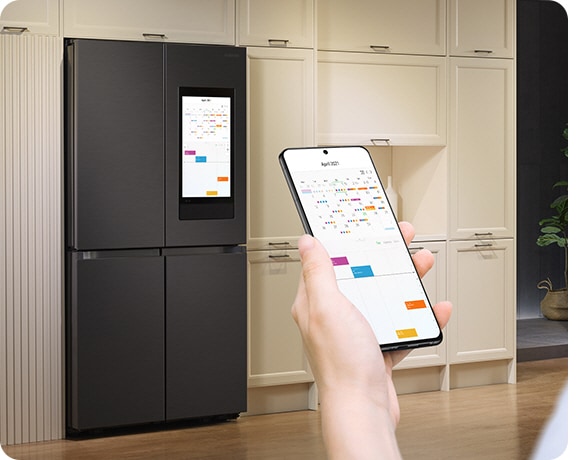 A hand holds a mobile phone with a calendar onscreen. The same calendar is on the Family Hub display in the background.