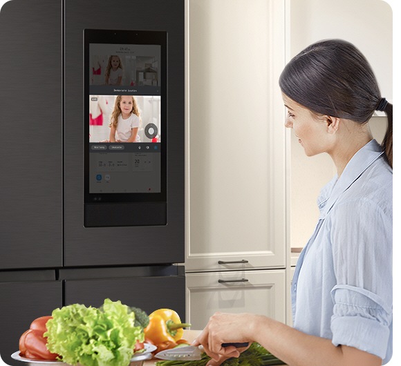 A woman chops veggies in a kitchen looking at the screen on a fridge, which shows a girl who is in another room.
