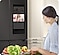 A woman chops veggies in a kitchen looking at the screen on a fridge, which shows a girl who is in another room.