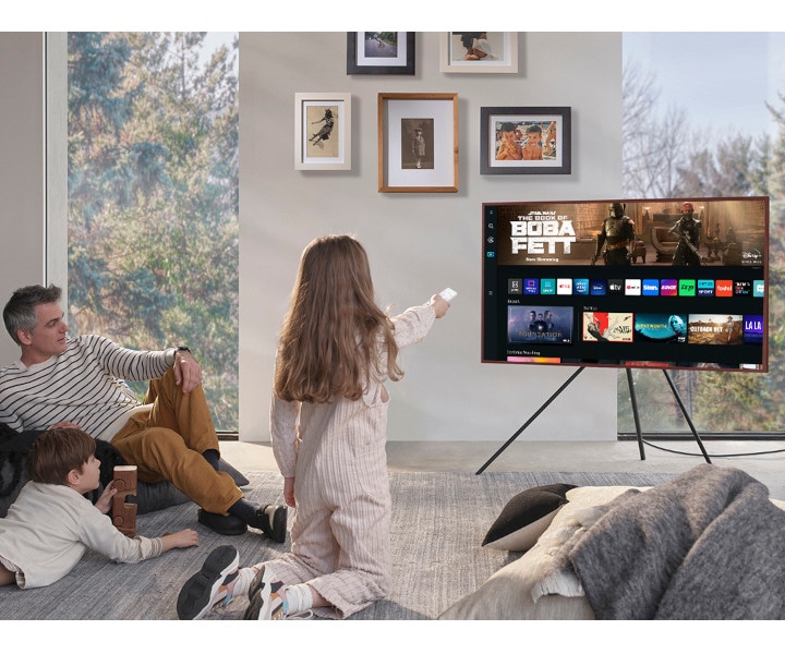 A girl is pointing the remote towards The Frame, which shows the Smart Hub home screen.