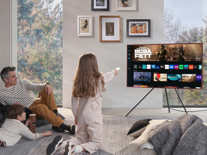 A girl is pointing the remote towards The Frame, which shows the Smart Hub home screen.