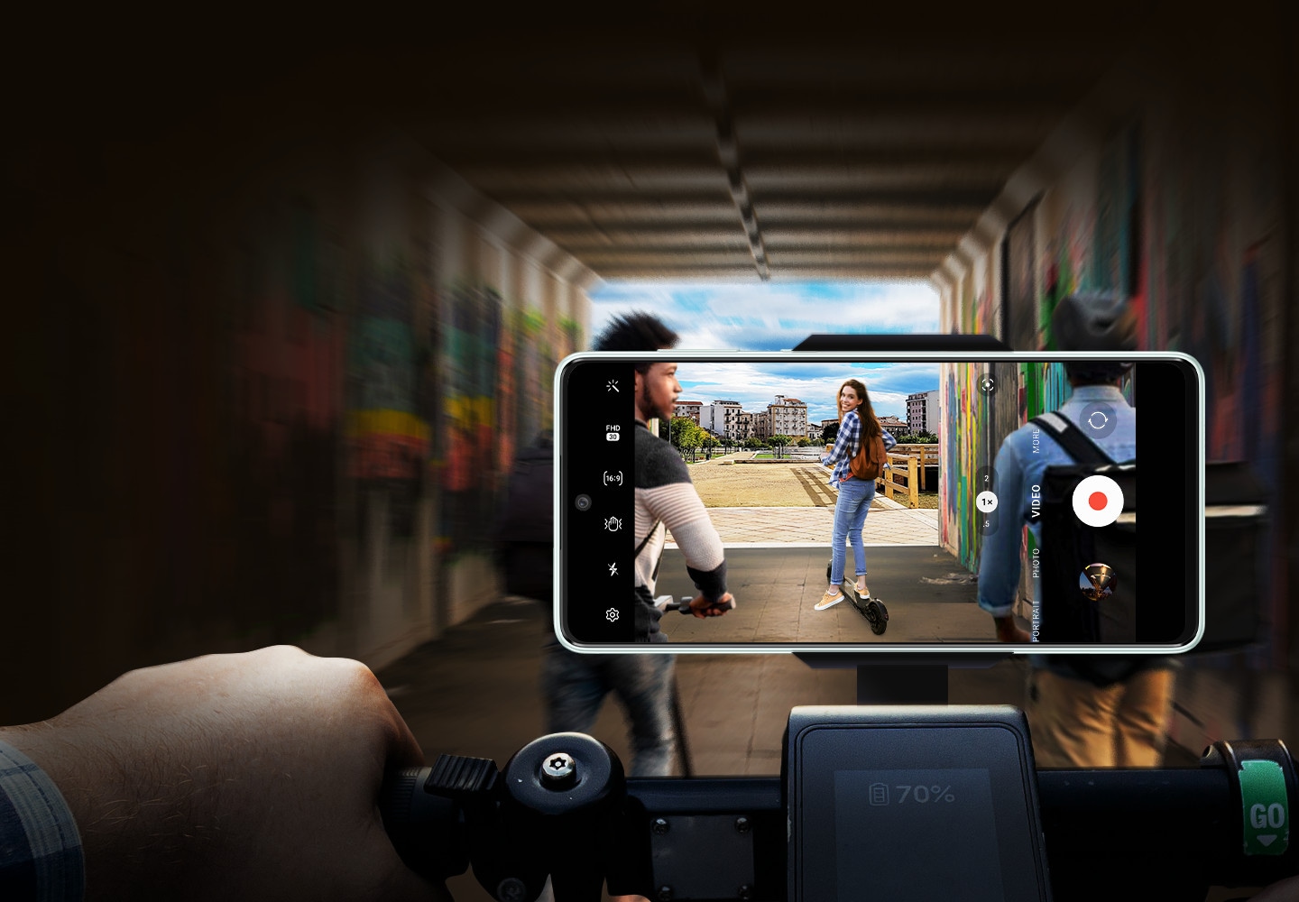 A Galaxy A73 5G device is mounted horizontally onto a bicycle handlebar and shooting a video. While the picture inside the frame clearly shows the electric kickboard riding up front and a woman in looking back at the device, the background image beyond the frame is dark and blurry.