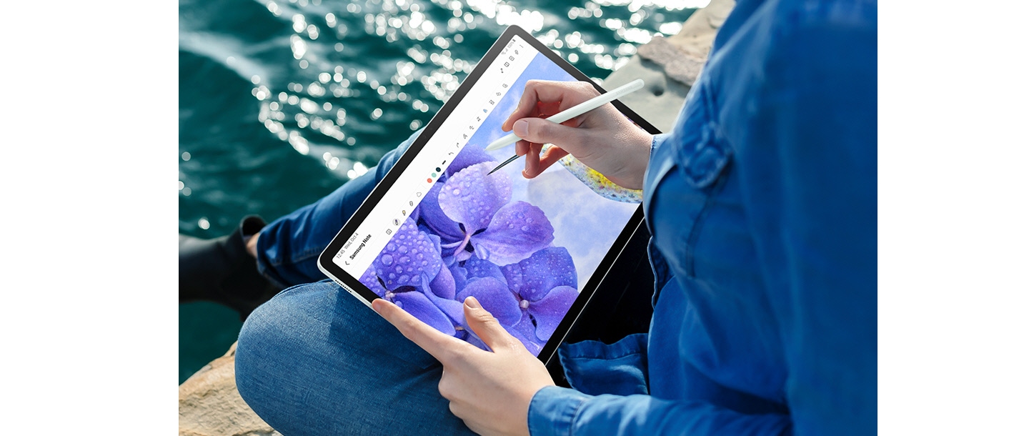 A person sitting near water is holding Galaxy Tab S9 FE+ in Landscape mode and using S Pen to draw a flower picture in the Samsung Notes app open onscreen.