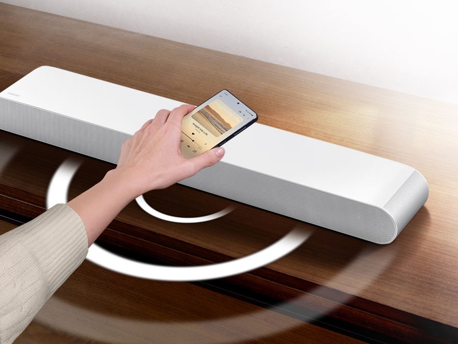 A hand holds a phone playing music and taps it lightly against a Soundbar. Instantly, the Soundbar gives off a series of round sound waves, indicating that it's playing the same music as the phone.