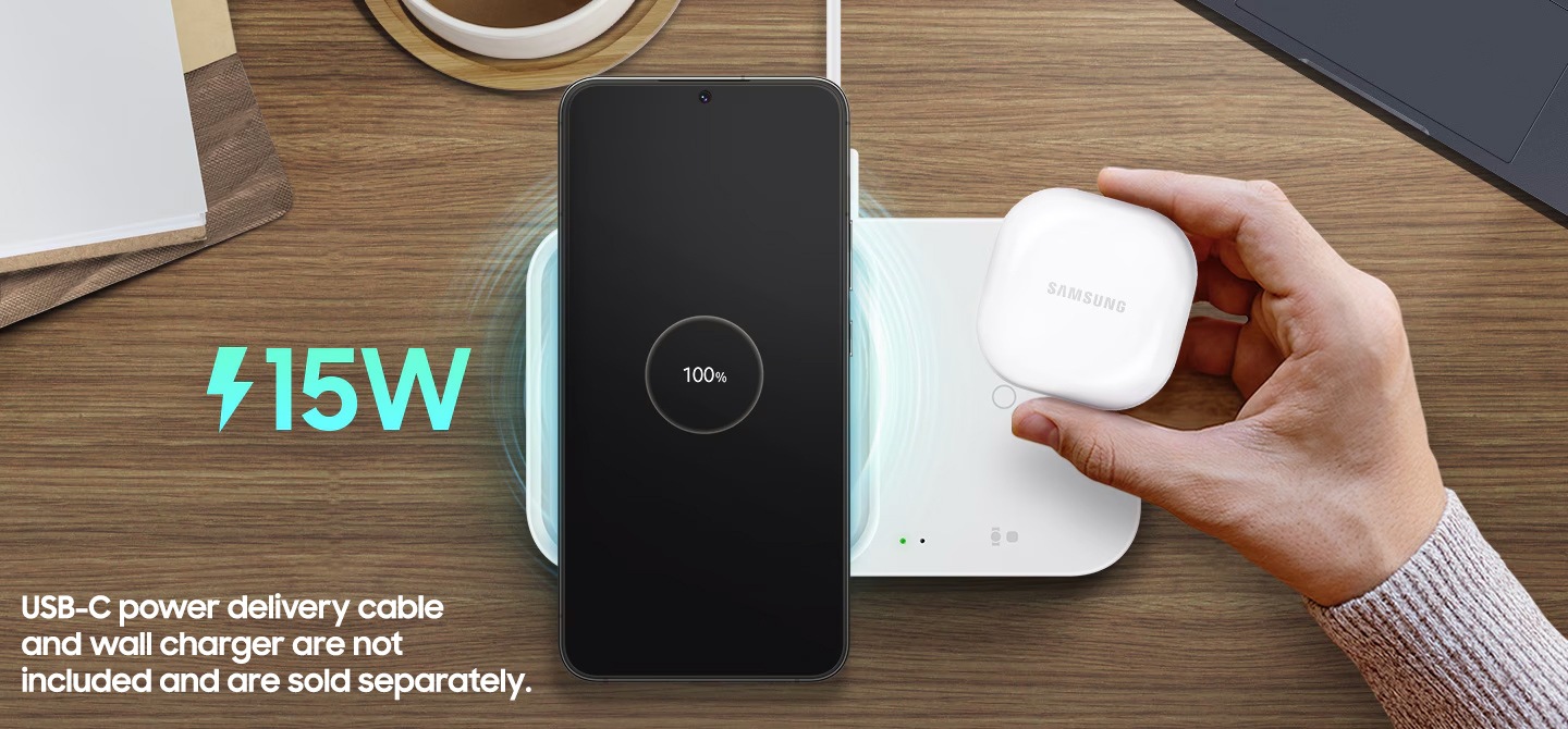 A 15W Wireless Charger Duo with power connected is on a wooden table. A black-colored Galaxy S22+ with the text 100% onscreen is on the left side of the unit. A hand is holding a white Galaxy Buds2 case to place it on the right side of the charger.