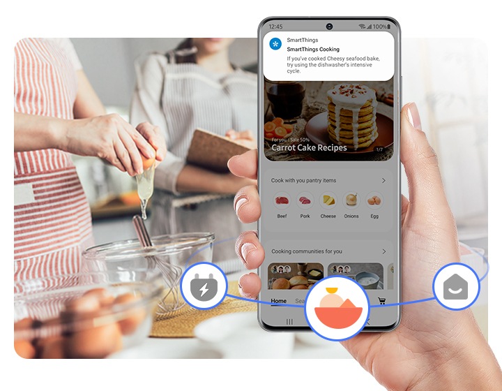 A hand holding a phone with a smartthings cooking alert displayed, with three smartthings icons visible in the surroundings: energy, cooking, and homecare, with the cooking icon highlighted. The background shows people cooking.