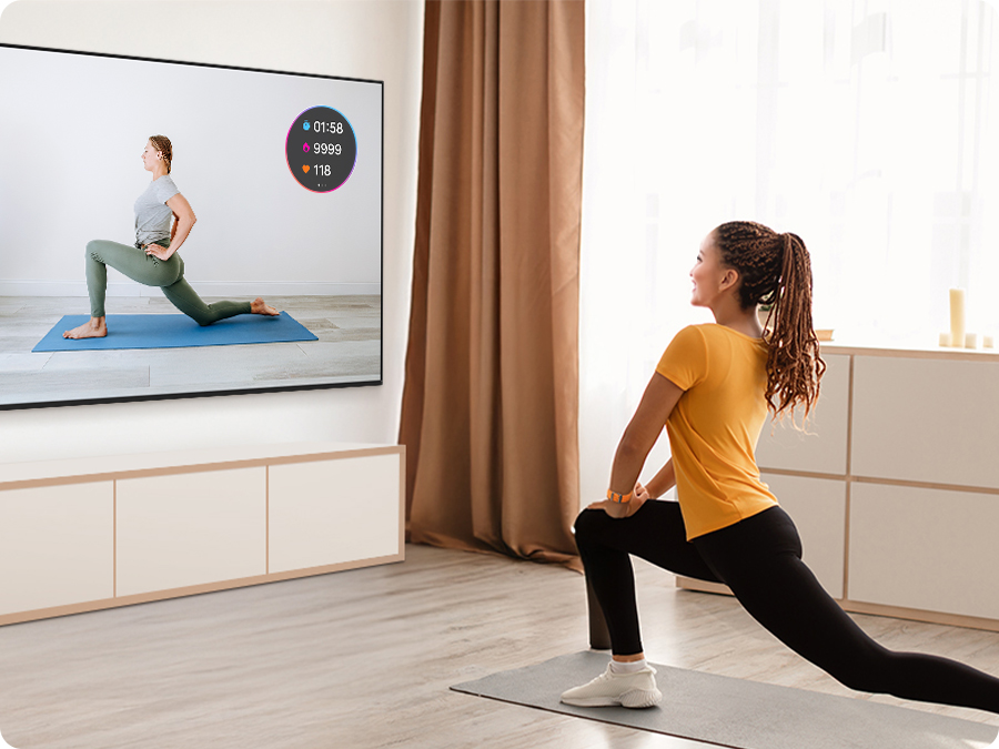 A woman exercises while watching a fitness trainer on TV. Her workout stats from Galaxy Watch appear as an on-screen overlay.