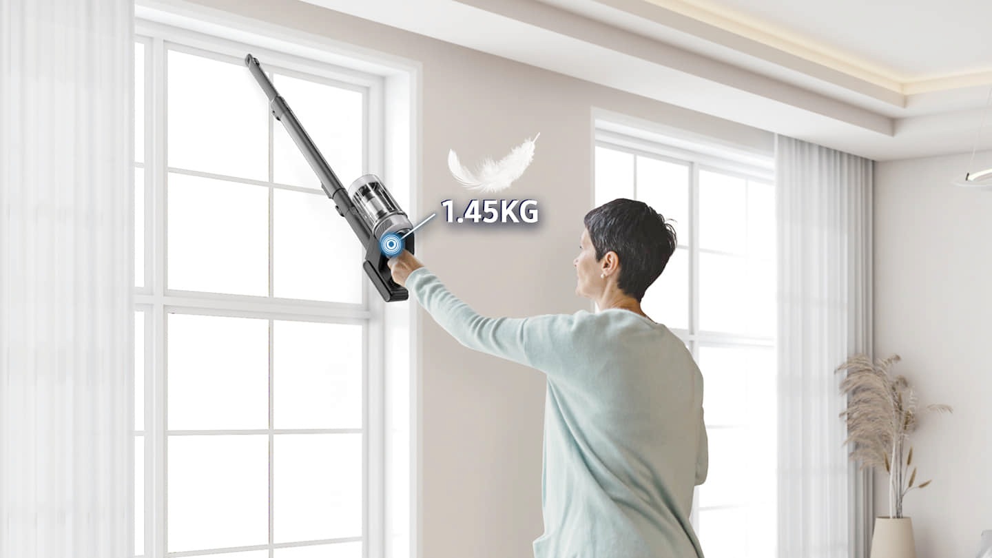 A person wearing a mint-colored top is cleaning a tall window with a lightweight 1.45kg cordless vacuum cleaner in a bright indoor space.