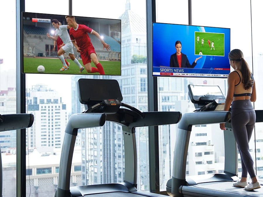 In a hotel gym with a view through large windows, a woman is walking on a treadmill. Each treadmill is equipped with a Hotel TV displaying sports content.