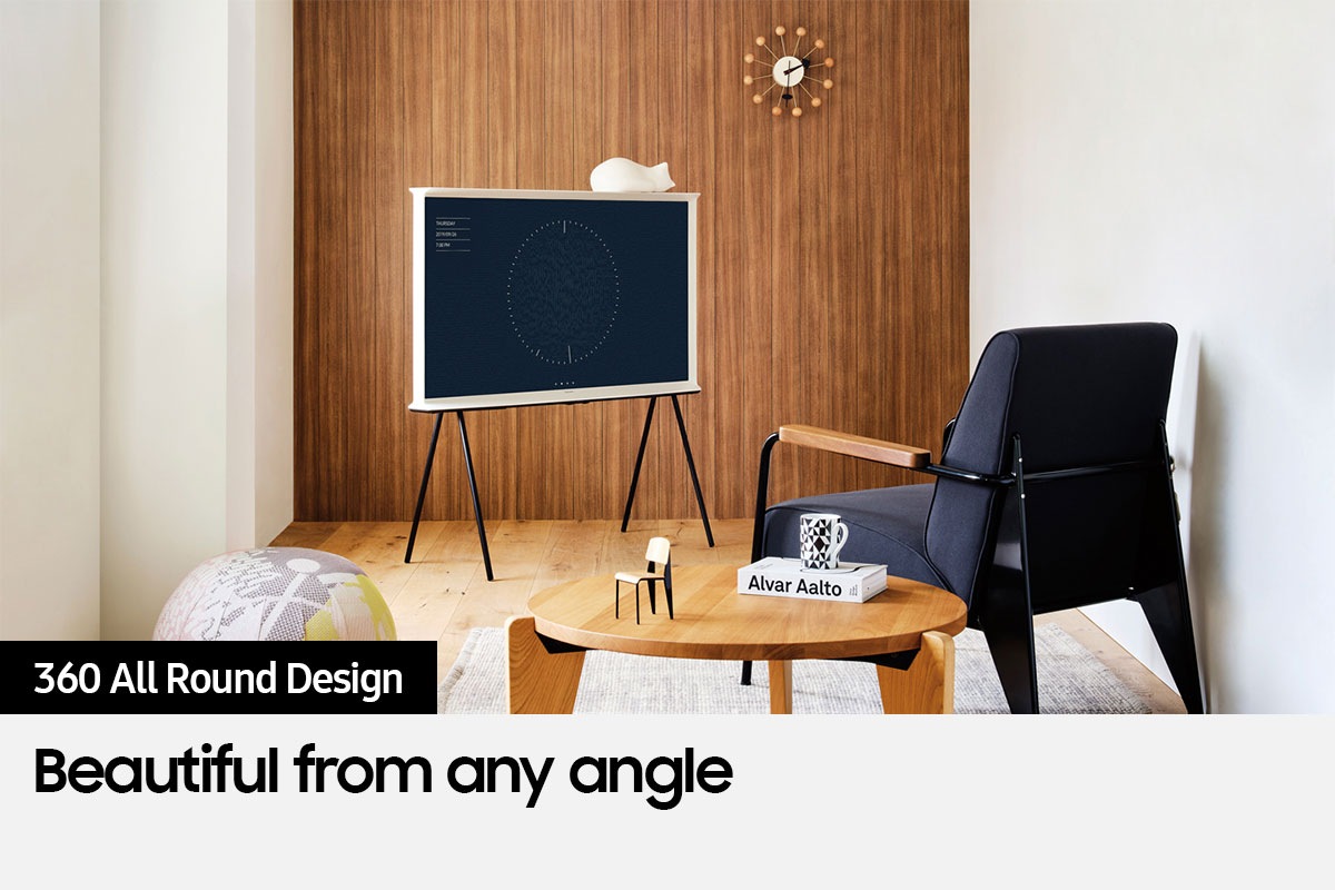 The Serif is on a stand next to a chair and small table.