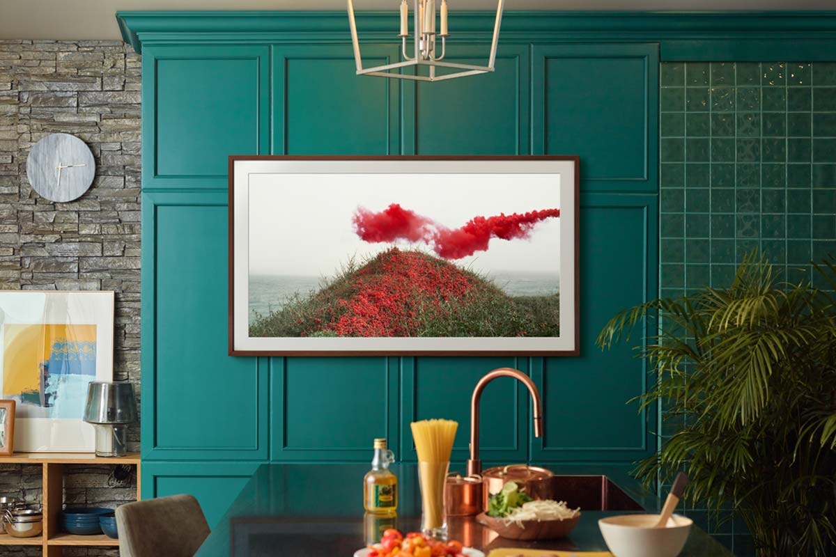 The Frame TV is wall-mounted using the Slim Fit Wall Mount accessory in a kitchen and is displaying a green landscape with red flowers and red smoke.