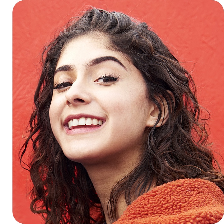 A selfie of a woman standing against a red wall that's gently blurred. She's clear, and seen smiling and looking down into the camera.