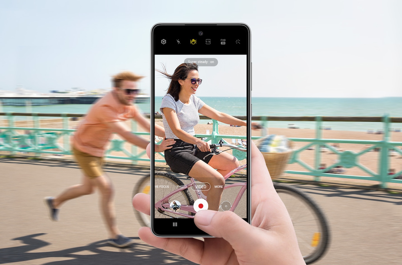 A person on a bike with another person running behind them and pushing. In front of this is a hand holding Galaxy A52s 5G with the camera interface onscreen. The scene in the display is clearer than the scene outside of the display, depicting how Super Steady allows video to be captured smoothly even if the subject is in motion.