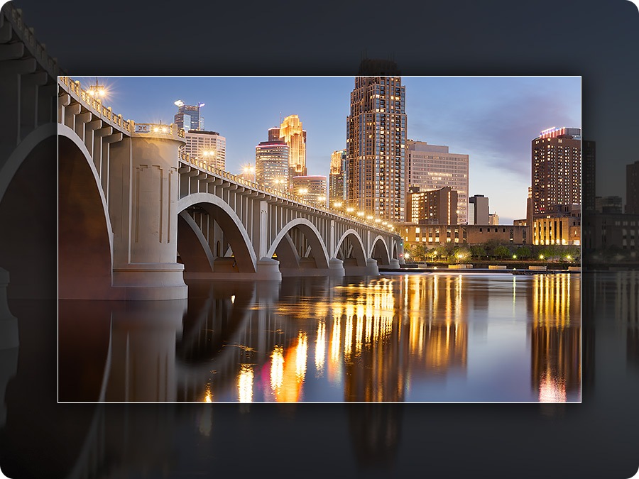 An image of a cityscape at twilight, where vibrant lights reflect beautifully on the calm water beneath a gracefully arched bridge.