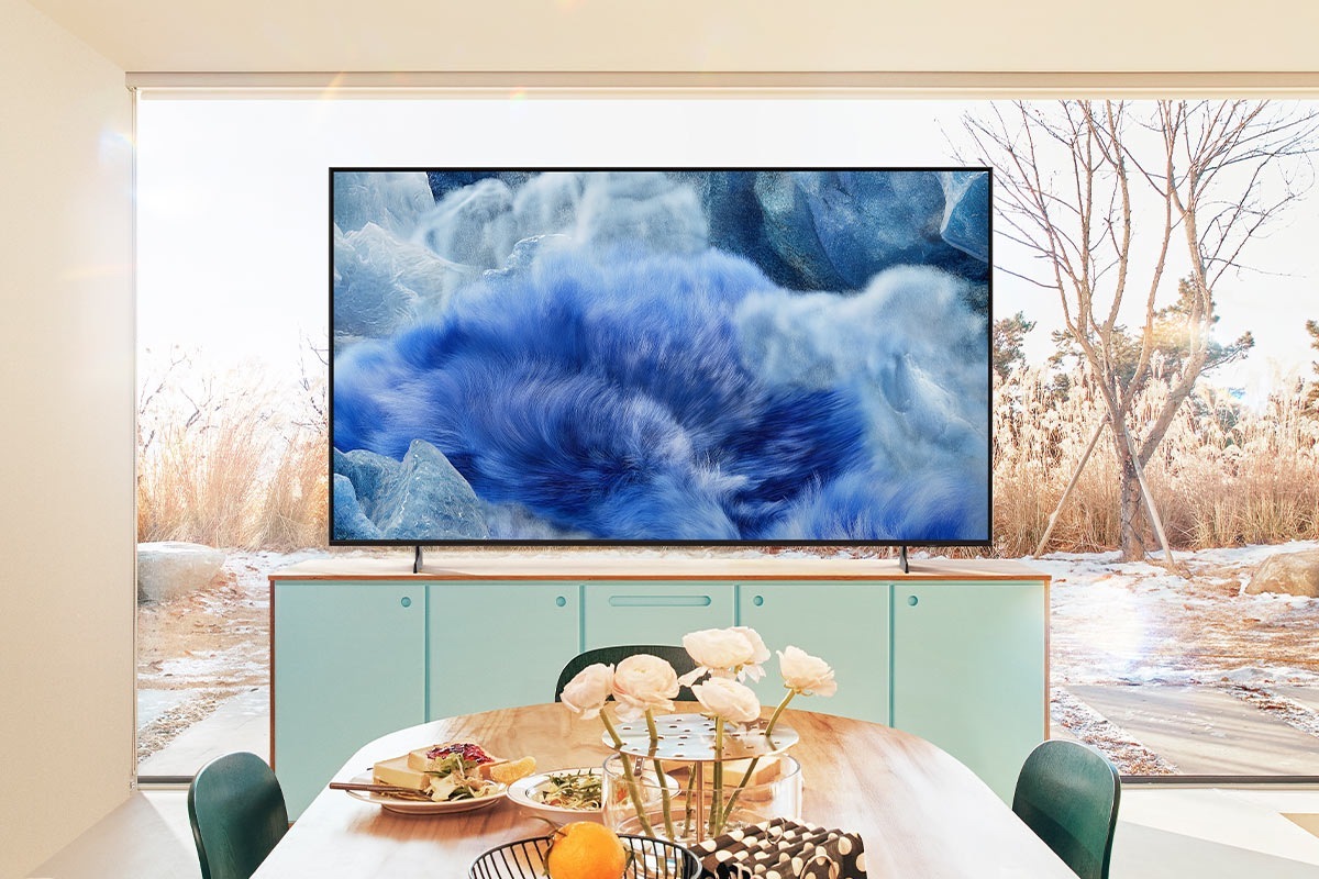 A QLED TV in a kitchen area displays a blue grassy landscape. Behind the TV, a glass window reveals the outdoor scenery.