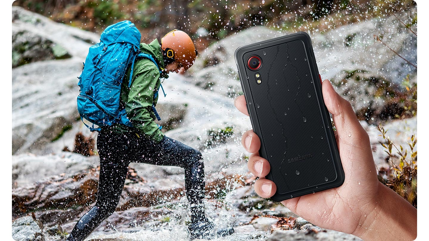 A hand is holding Galaxy XCover 5, showing backside with water droplets. In background, a person wearing orange helmet with blue backpack is crossing over the river, splashing water.