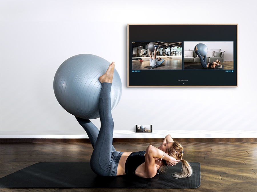 Una mujer está haciendo ejercicio con una pelota ejemplos en el grupo con su teléfono inteligente a su lado. El marco muestra lo que hay en su teléfono inteligente, así como un video de un entrenador en la pantalla del televisor