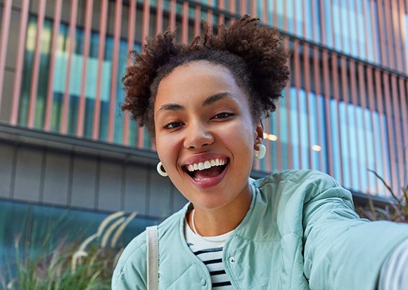 Un selfie d'une femme souriante avec un bâtiment moderne à l'arrière-plan.