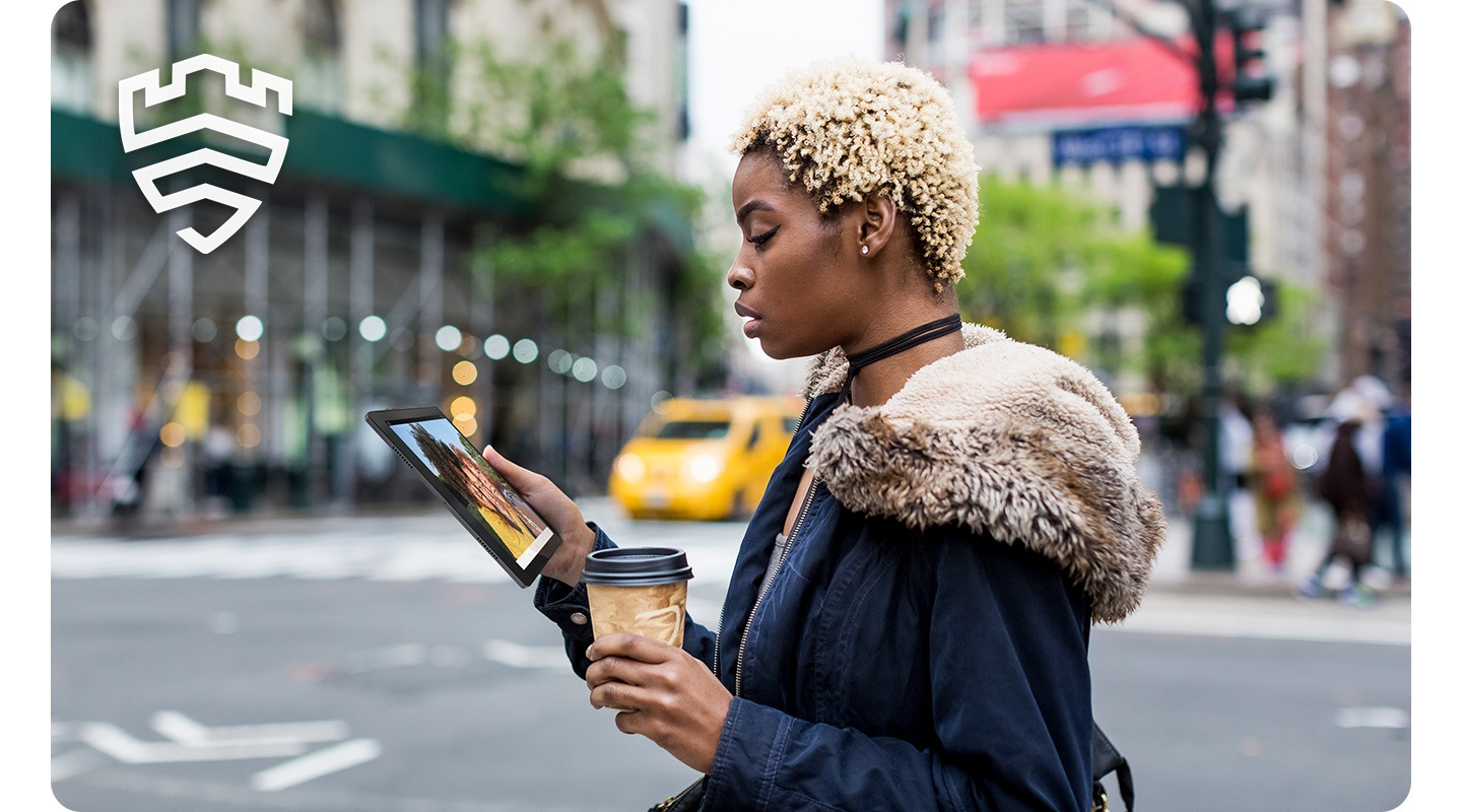 Mulher segurando um dispositivo Galaxy Tab A8 em uma das mãos e uma bebida para viagem na outra olha para a tela em uma rua da cidade.