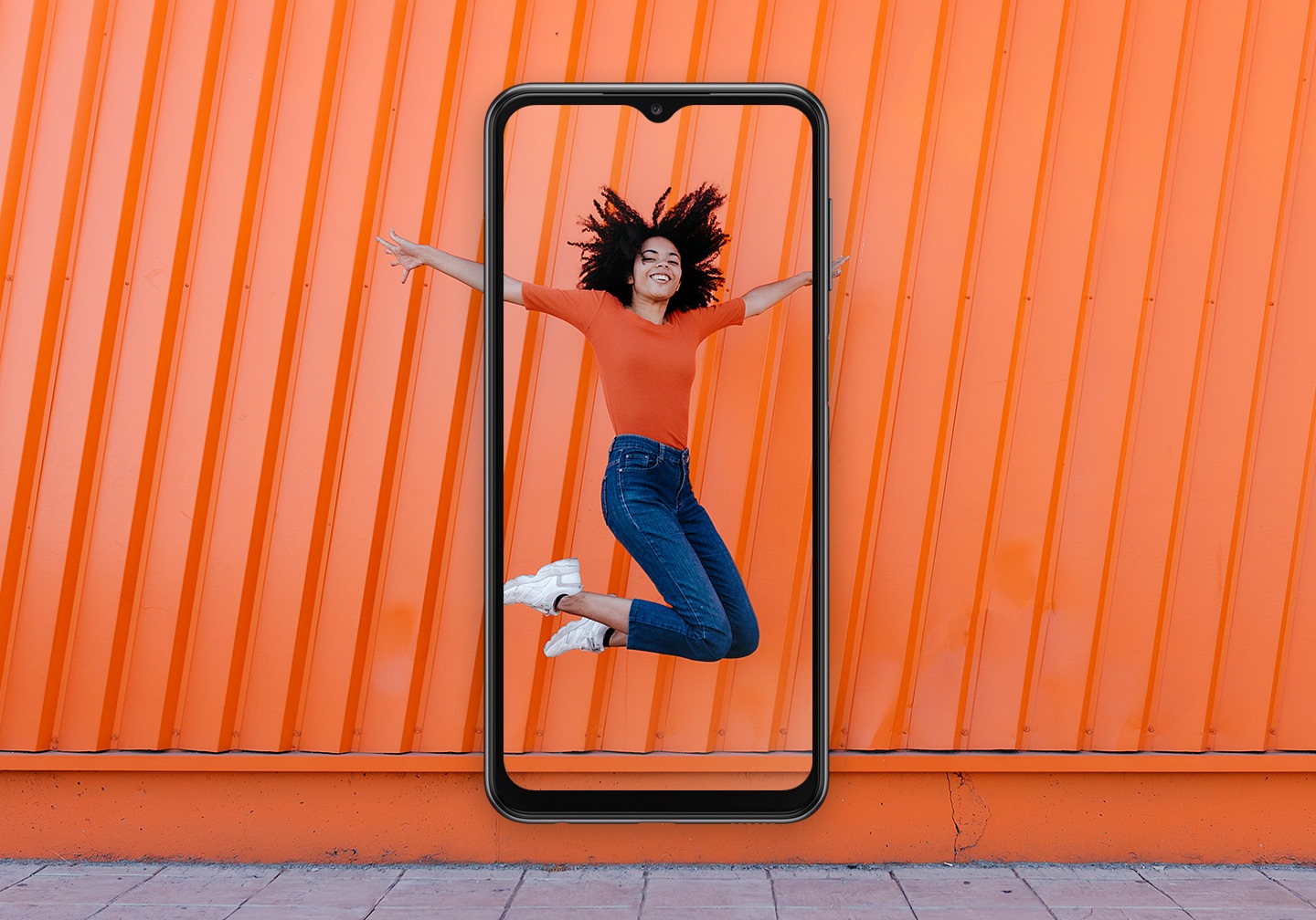  Uma mulher, sorrindo, está saltando em frente a um fundo de parede laranja. No centro, um Galaxy A23 está se sobropondo e captura a mulher dentro da tela.